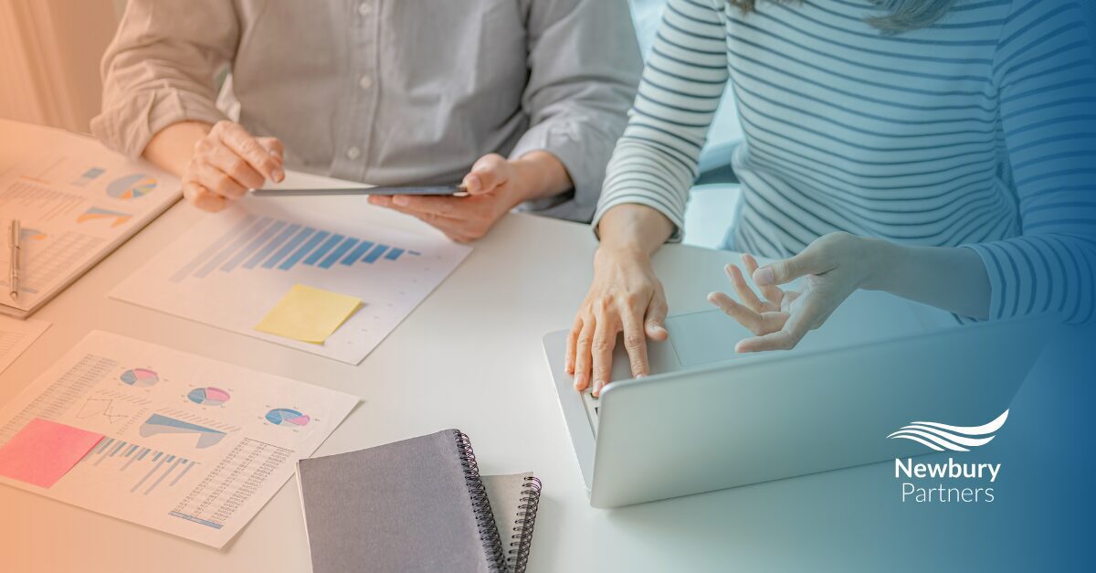Two people collaborate on a laptop at a table, surrounded by papers and graphs, focusing on staffing tech stack optimization.